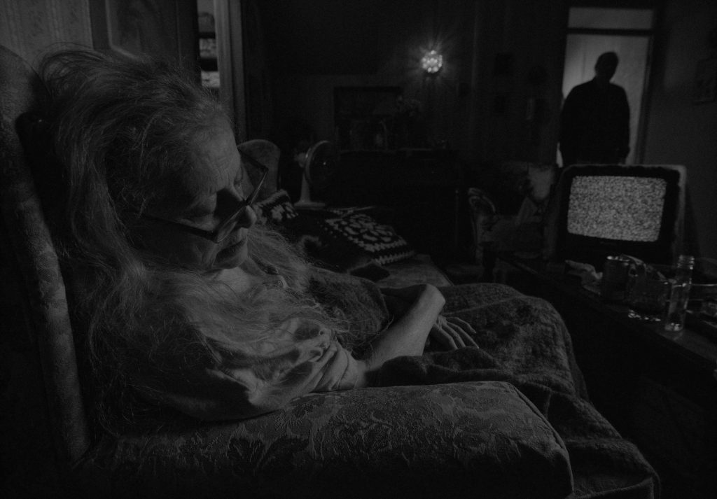 screenshot di uno dei cortii di Deep Night,, in bianco e nero, raffigurante una signora che dorme in salotto, sulla porta davanti a lei una figura inquietante