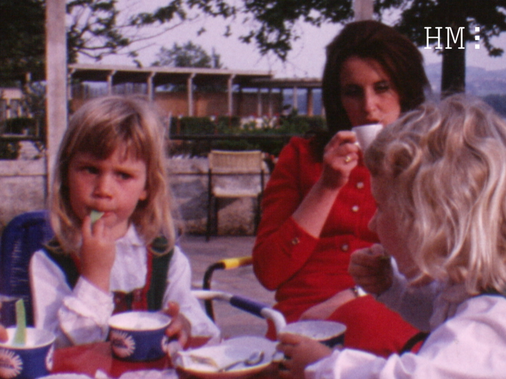 Fotogramma di filmato di archivio raffigurante una donna vestita di rosso che beve un caffè, alla sua sinistra una bambina bionda che mangia un gelato, di fronte a loro, un'altra bambina bionda che ci dà le spalle